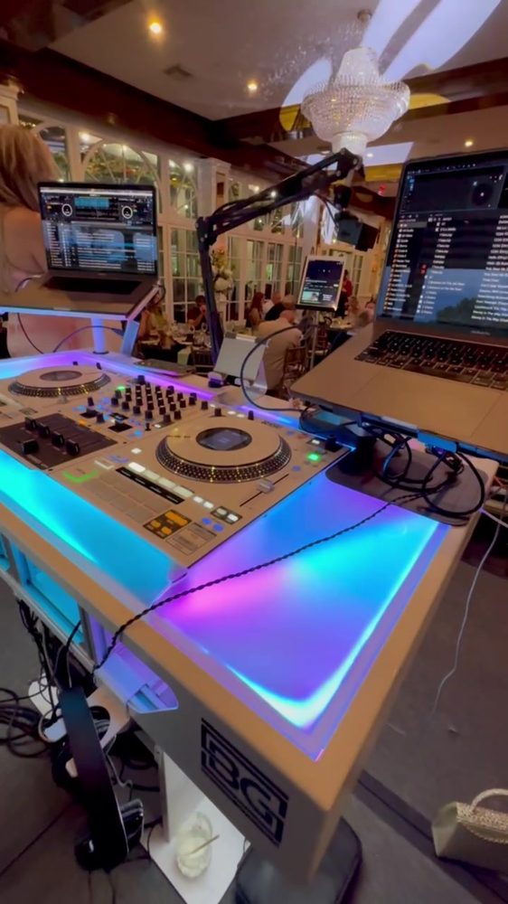 The all-white DJ booth with its acrylic top glowing cyan and magenta under a white controller, a chandelier behind it